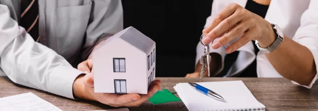 A person hands over keys to another person across a table with a model house, pen, and documents, indicating a real estate transaction.