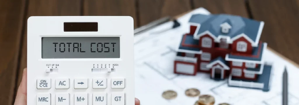 Calculator displaying 'Total Cost' in focus, with a model house, blueprints, and coins blurred in the background.