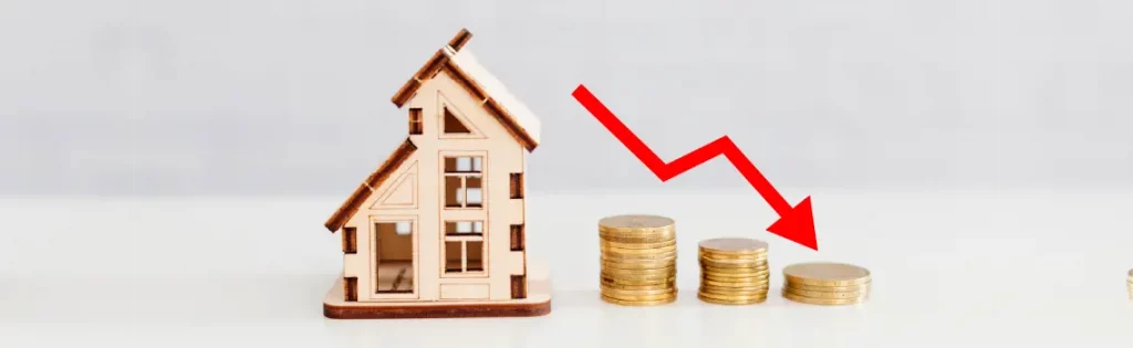 House model next to stacked coins with a red downward arrow, symbolizing declining home prices or value.