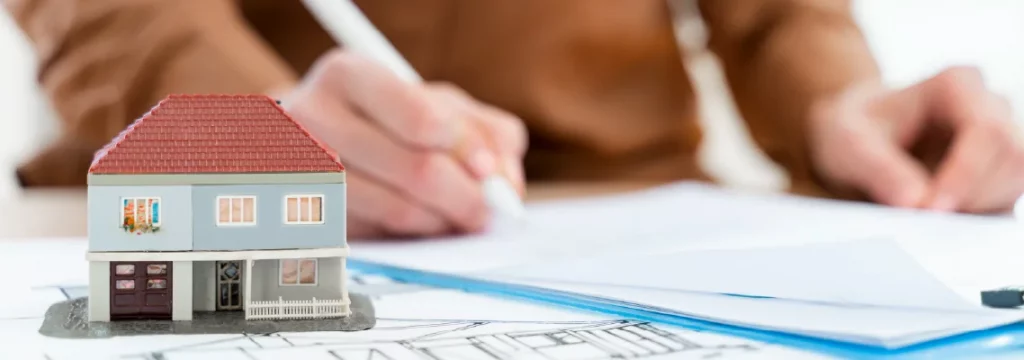 Small model house placed on blueprints with a person in the background reviewing documents and taking notes.