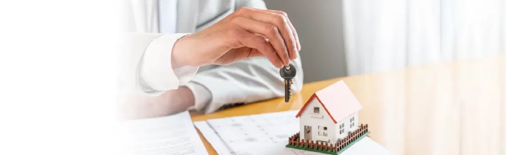 Person holding house keys above a small house model on a desk, symbolizing a real estate purchase or mortgage.