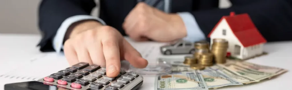 Person using a calculator with cash, coins, and a house model nearby, symbolizing mortgage or financial planning.