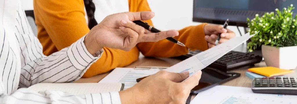 Two professionals collaborate at a desk, examining financial documents with calculators and pens in hand.