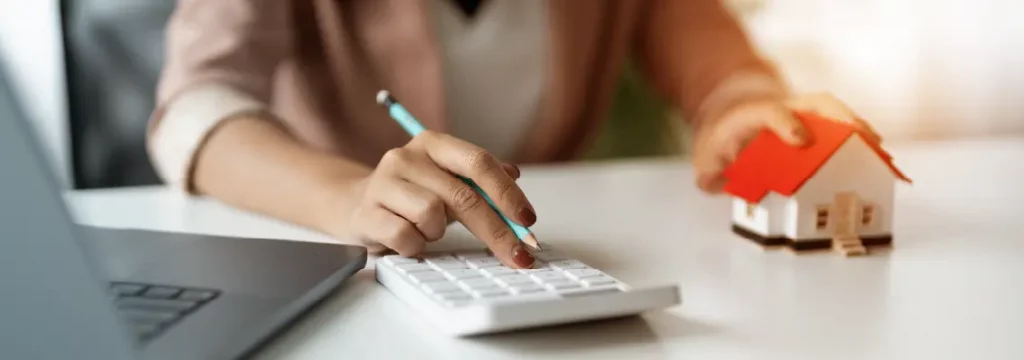 Real estate agent calculates finances on a calculator while positioning a model house on the desk.