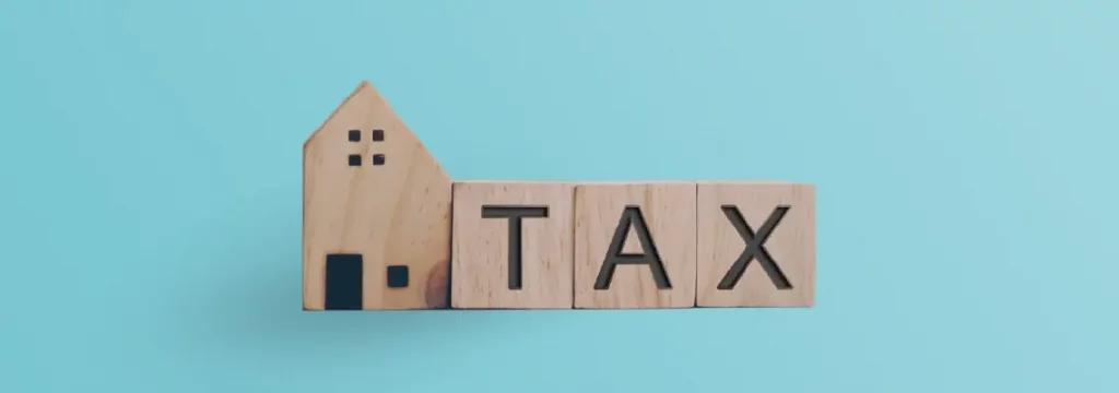 Wooden blocks spelling "TAX" beside a simplistic model house, all set against a vivid blue background.