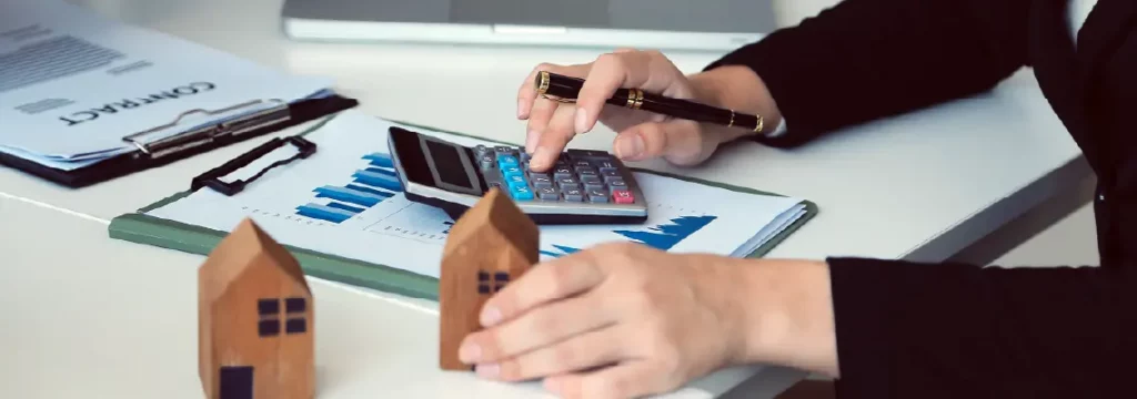 Financial expert calculates figures on a calculator, with charts and model homes on the desk.