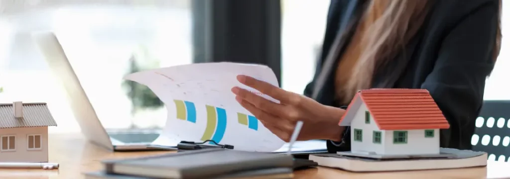 Real estate professional reviews financial charts with laptop and scale model homes on her desk.