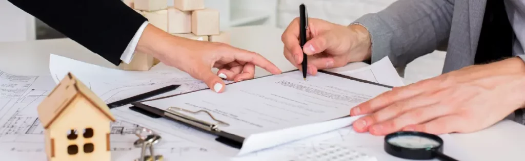 Hands signing mortgage documents with house model and keys nearby, representing a home purchase.